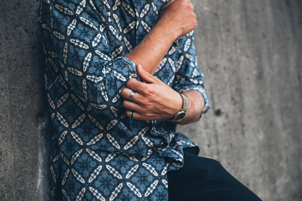 25-30 year old,adult,blue,clothing,grey,hand,one person,portrait,standing,arms,caucasian,concrete walls,fashion,fashionable,leans on wall,lifestyle,long hair,male,man,outside,patterned shirt,person,rings,style,stylish,wrist watch