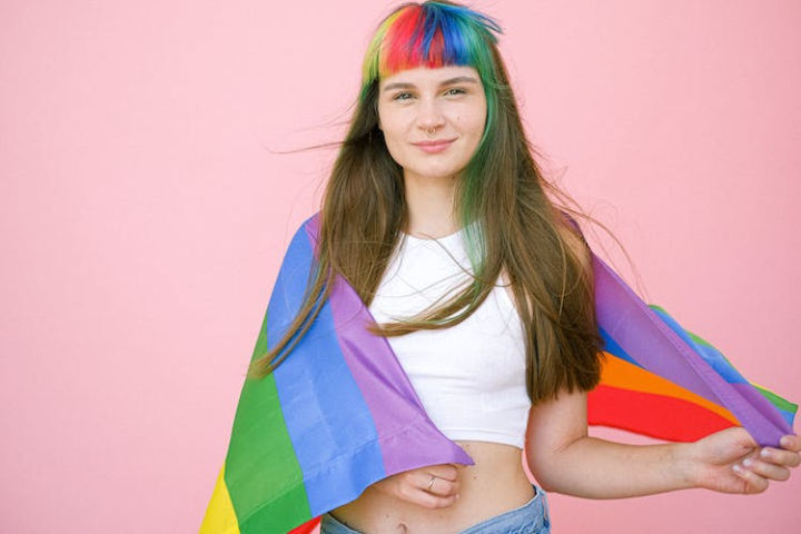 colored hair,copy space,cute,gay pride,gay pride flag,hair,hairstyle,holding,lgbt,lgbt pride,lgbtq,looking at camera,model,pink background,pretty,rainbow,rainbow bangs,rainbow flag,smile,woman,young woman