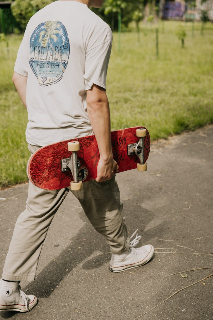 back view,blurred background,carrying,holding,park,paved pathway,person,selective focus,skateboard,skateboarder,skater,vertical shot,walking,young