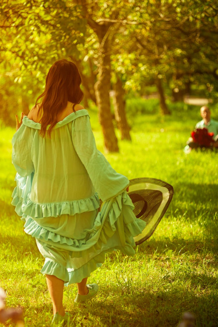 back view,blurred background,grass,green dress,outdoors,sun hat,trees,vertical shot,walking,woman