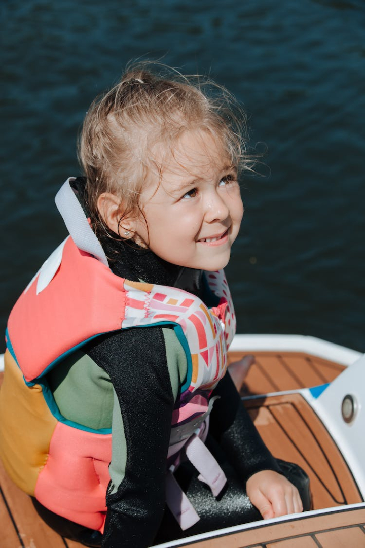 blond hair,boat,girl,high angle view,human face,kid,long hair,looking up,outdoors,sitting,vest,water