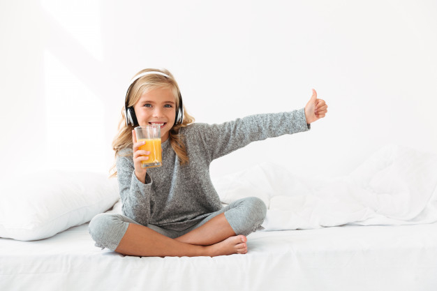 toothy,offspring,indoors,crossed,showing,little,cheerful,daughter,small,bedding,blonde,pajamas,childhood,pretty,listening,holding,nursery,beverage,legs,soft,thumb,sitting,fresh,bedroom,morning,headphones,bed,finger,juice,glass,room,child,kid,happy,home,girl,music
